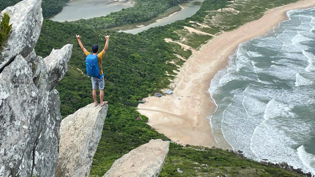 Reisender mit Rucksack mit gereckten Armen auf Felsen hoch über einem Strand