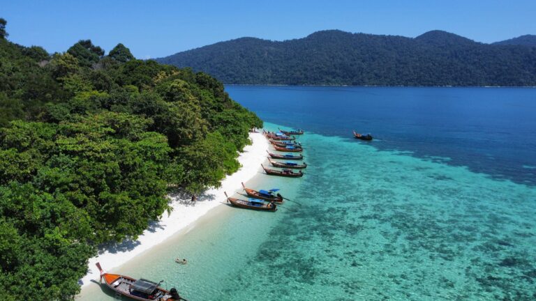 Luftaufnahme eines weißen Sandstrandes mit traditionellen Longtailbooten, türkisblauem Meer und dichtem tropischem Regenwald in Thailand