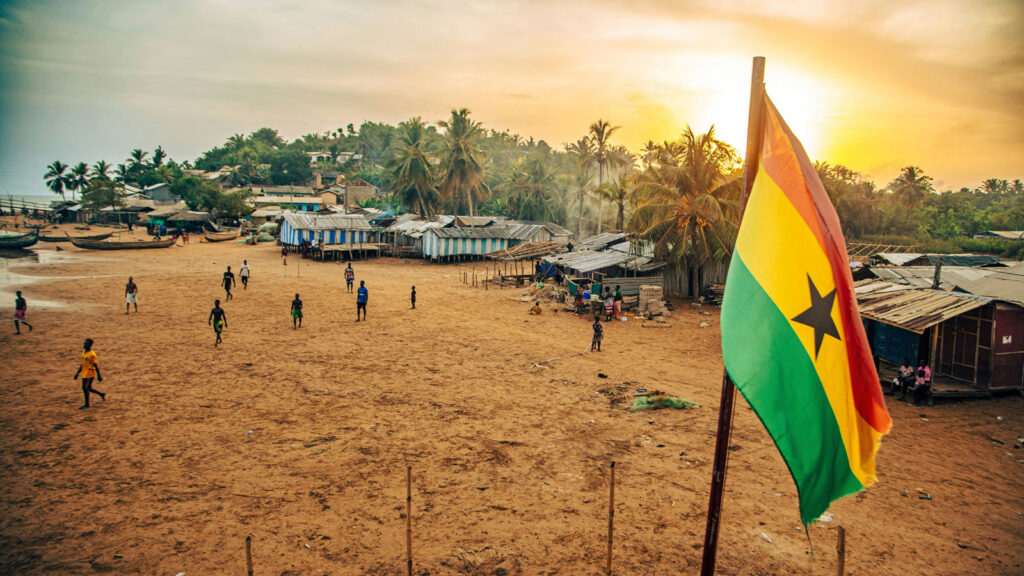Ghana Flagge vor Strand mit Häusern und Menschen bei Sonnenuntergang
