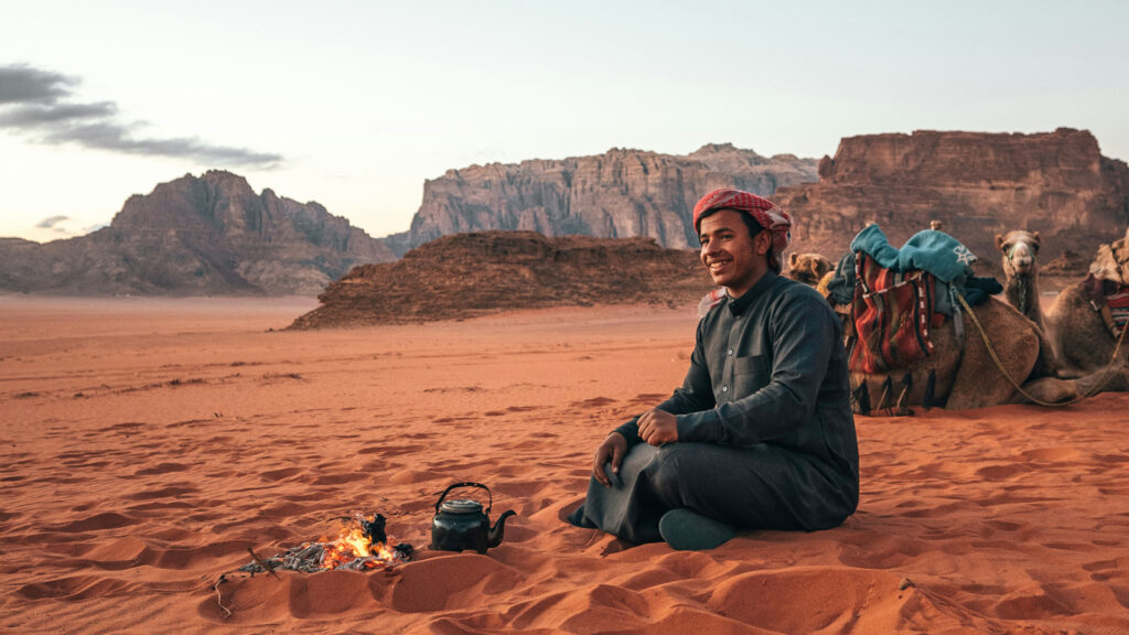 Traditionell gekleideter Mann vor Lagerfeuer mit Teekessel und Kamel in der Wüste in Wadi Rum