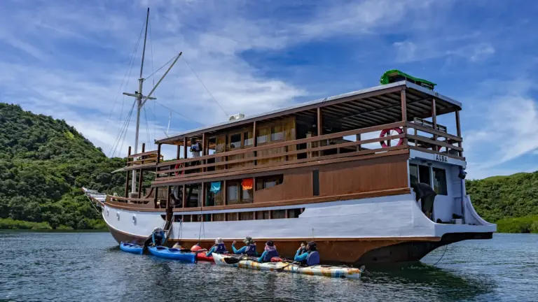 Traditionelles Holzboot vor tropischer Inselkulisse in Indonesien mit Kajakfahrern – Inselhopping Reise durch Bali, Lombok, Gili und Flores