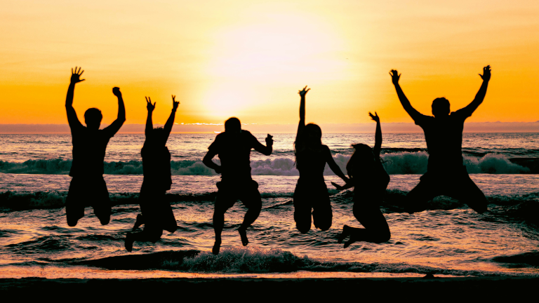 Junge Reisende springen gemeinsam bei Sonnenuntergang am Strand und erleben Gemeinschaft auf einer Gruppenreise für junge Leute