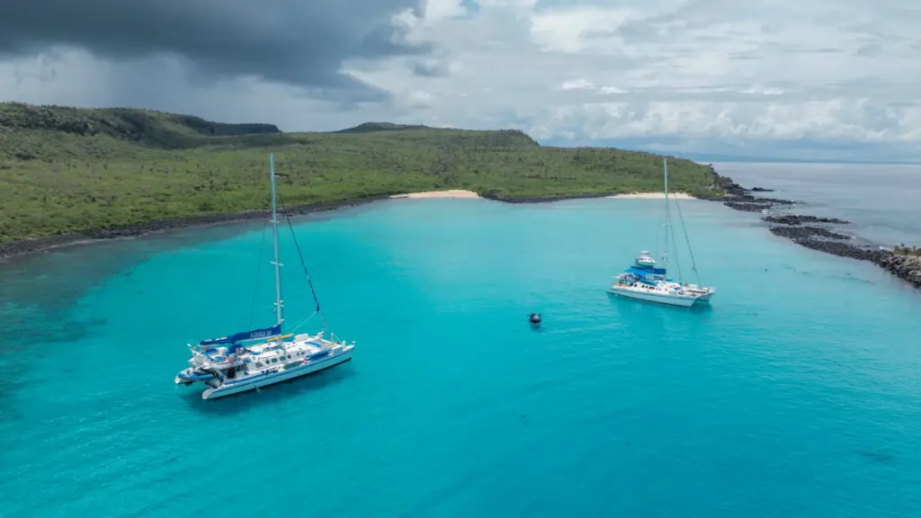 Katamaran Nemo III und ein weiteres Segelboot ankern in einer türkisblauen Bucht der Galapagos-Inseln vor einer grünen Vulkanlandschaft
